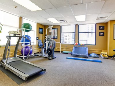 Open rehabilitation gym with a treadmill and conditioning equipment beside tall windows.