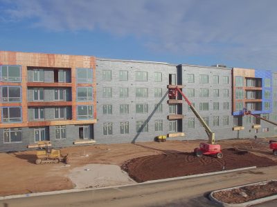 Four-story modern loft-style apartment building under construction, with grey and wood-tone facade panels and active site work.