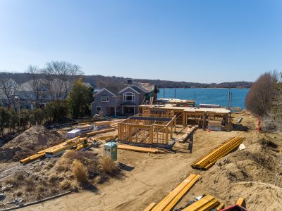 Framed waterfront home under construction with stacked lumber and a bay view in Northport, New York.