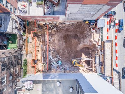 Top-down aerial of a tight urban building site with an excavated foundation, safety barriers, and construction vehicles.